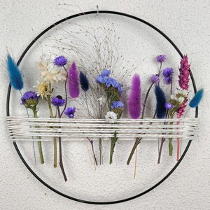 Op de afbeelding: Een zwarte metalen ring met gedroogde bloemen en grassen die erin zijn geweven. De bloemen zijn in tinten paars, blauw, wit en roze. De ring hangt aan een witte muur.