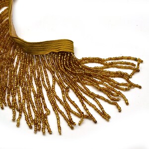 May include: Gold beaded fringe trim with a woven gold band. The fringe is made of small, shiny beads, creating a textured and decorative element. Ideal for embellishing clothing, accessories, or home decor projects.