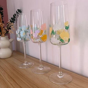 May include: Three champagne flutes with hand-painted floral designs. The flutes are clear glass and feature different coloured flowers, including blue, pink, yellow, and white.