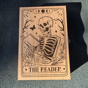 May include: Wooden tarot card featuring a black illustration of a skeleton reading a book, with floral and celestial accents. The text "THE READER" is at the bottom. The card has a light brown wood grain.