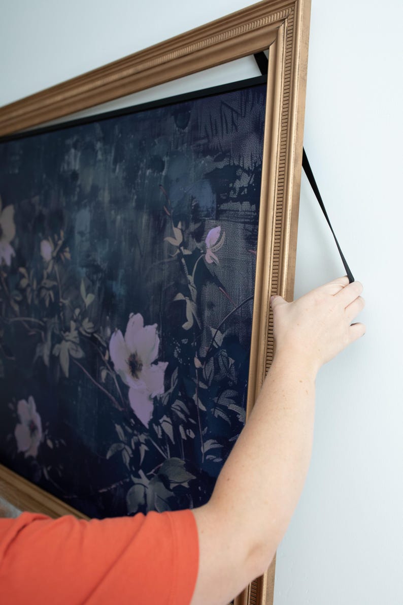 以下が含まれることがあります： ダークネイビーの背景に淡いライトパープルの花柄が描かれた長方形のウォールアート。質感のあるゴールドトーンのフレームに収められており、フレーム上部に取り付けられた黒いストラップで吊り下げられています。