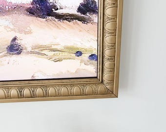 May include: A framed oil painting featuring a landscape with textured brushstrokes in shades of cream, beige, and deep purple. The artwork is encased in an ornate, metallic gold frame with a repeating arched pattern along the inner border.