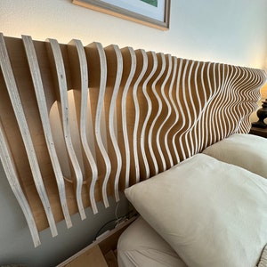 Op de afbeelding: Een modern houten hoofdbord met een uniek golfachtig ontwerp. Het hoofdbord is gemaakt van lichtgekleurd hout met verticale, gebogen lamellen. Het ontwerp omvat ingebouwde verlichting langs de bovenrand, waardoor een zachte gloed ontstaat. Het hoofdbord bevindt zich boven een bed met wit beddengoed.