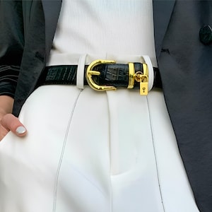 May include: Black leather belt with a gold-colored buckle and a small gold-colored padlock. The belt has a crocodile skin texture and is worn with white pants and a black blazer.