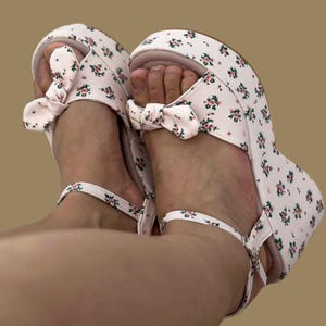 Op de afbeelding: Paar plateausandalen met een bloemenprint in roze en groen op een witte achtergrond. De sandalen hebben een strikdetail aan de voorkant en enkelbandjes. De sleehak biedt lift en ondersteuning.