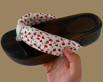 May include: Wooden platform sandal with a floral fabric strap. The dark brown wood base has a curved shape with a small brown pad for the foot. The strap is white with a pattern of small red flowers and gray leaves.