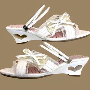 May include: White wedge sandals with heart cutouts, bows, and lace detailing. The sandals have a strap that goes around the ankle and a decorative bow on the top. The heels are approximately 3 inches high.