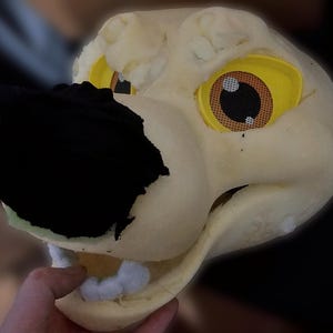 May include: Unfinished white foam animal head with black fabric nose and yellow and orange eyes. The head is being held by a hand.