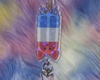 May include: A clear acrylic keychain featuring a red, white, and blue popsicle with a cartoon fox face. The popsicle is dripping blue liquid and has white stars on it. The keychain has a silver star-shaped charm.
