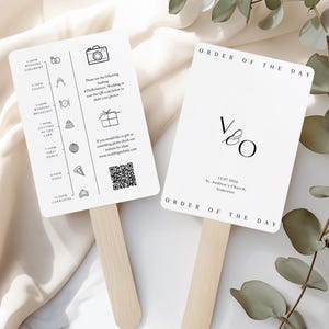 Può includere: Due ventagli da matrimonio bianchi con manici in legno. Un ventaglio mostra un programma con icone e testo, incluso un codice QR. L'altro ventaglio ha le iniziali "V & O" e la data "13.07.2026". Entrambi i ventagli riportano "Ordine del giorno".