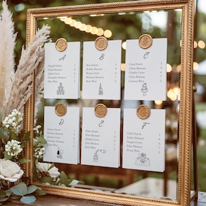 May include: A gold framed seating chart with six white cards, each held by a gold wax seal. The cards display guest names and table numbers, with decorative illustrations. The frame is set on a wooden surface, with floral arrangements in the foreground.
