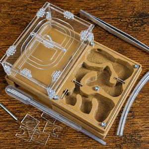 May include: A tan ant farm with a clear acrylic lid and a multi-chambered interior. The set includes metal tweezers, a clear plastic pipette, a flexible tube, a small L-shaped metal tool, and several small clear acrylic dividers for the chambers.