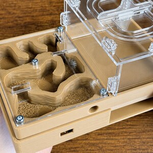 May include: A tan and clear acrylic ant farm with a maze-like interior. The farm has a clear top and is held by a person's hand. The interior has a sandy texture, and the top is secured with small screws.