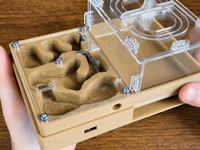 May include: A tan and clear acrylic ant farm with a maze-like interior. The farm has a clear top and is held by a person's hand. The interior has a sandy texture, and the top is secured with small screws.