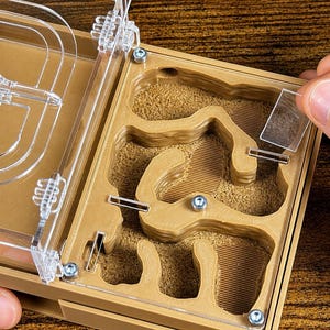 May include: A tan and clear ant farm with a clear plastic lid. The farm has multiple chambers and tunnels filled with a sandy material. A person's hand is placing a clear plastic piece into the farm.