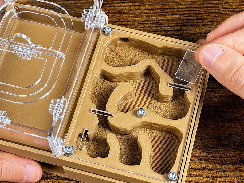 May include: A tan and clear ant farm with a clear plastic lid. The farm has multiple chambers and tunnels filled with a sandy material. A person's hand is placing a clear plastic piece into the farm.