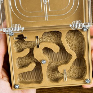 May include: A tan ant farm with a clear plastic lid. The interior features a complex network of tunnels and chambers, with a textured, sandy substrate. The lid is secured with small, silver screws. The overall design is functional and educational.