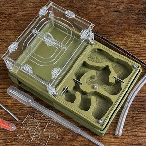 May include: An olive green ant farm with a clear acrylic top and a detailed interior. Includes a pipette, a small vial with red contents, a metal tool, and clear tubing. The farm is designed for ant observation and habitat.