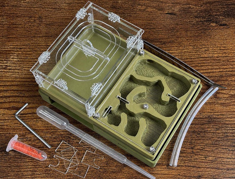 May include: An olive green ant farm with a clear acrylic top and a detailed interior. Includes a pipette, a small vial with red contents, a metal tool, and clear tubing. The farm is designed for ant observation and habitat.