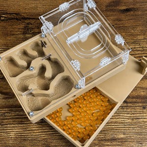 May include: A clear acrylic and tan ant farm with a removable lid. The farm has a built-in nest area with tunnels and chambers. A tray filled with orange spheres is visible. The ant farm is designed for ant observation and habitat.