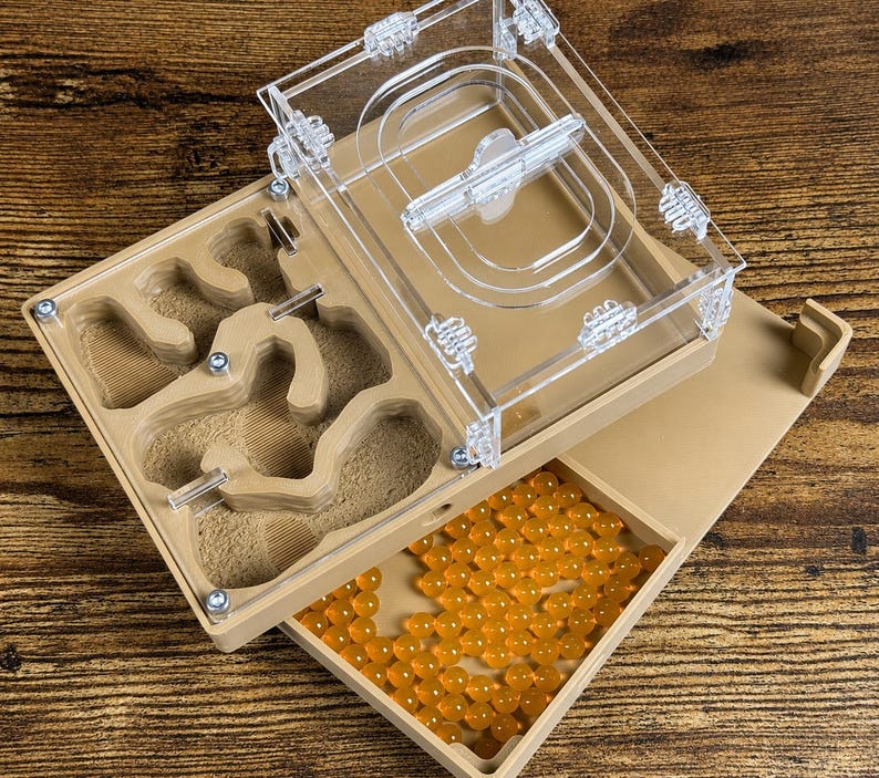 May include: A clear acrylic and tan ant farm with a removable lid. The farm has a built-in nest area with tunnels and chambers. A tray filled with orange spheres is visible. The ant farm is designed for ant observation and habitat.