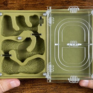 May include: An open, olive-green ant farm with a clear acrylic lid. The interior features a complex network of tunnels and chambers. The lid is attached with clear hinges and clasps. The farm is held by two hands.