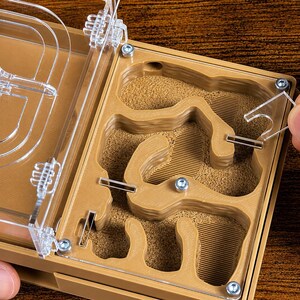 May include: A tan ant farm with a clear plastic lid is shown open. The interior has a sand-like substrate and carved tunnels. A hand is placing a clear plastic piece into the farm. The farm is on a wooden surface.