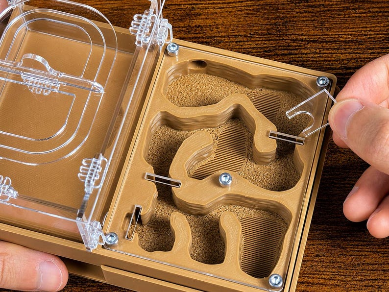 May include: A tan ant farm with a clear plastic lid is shown open. The interior has a sand-like substrate and carved tunnels. A hand is placing a clear plastic piece into the farm. The farm is on a wooden surface.