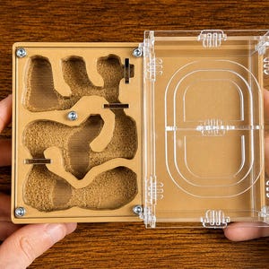 May include: An open, clear acrylic ant farm with a tan interior. The interior has a network of tunnels and chambers filled with a sandy material. The hinged lid is also clear, with a series of curved lines. The ant farm is held by two hands.