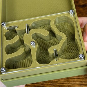 May include: A green ant farm with a clear top, revealing a maze-like interior filled with a textured, green material. The farm is held open, showcasing the design and construction with visible screws.