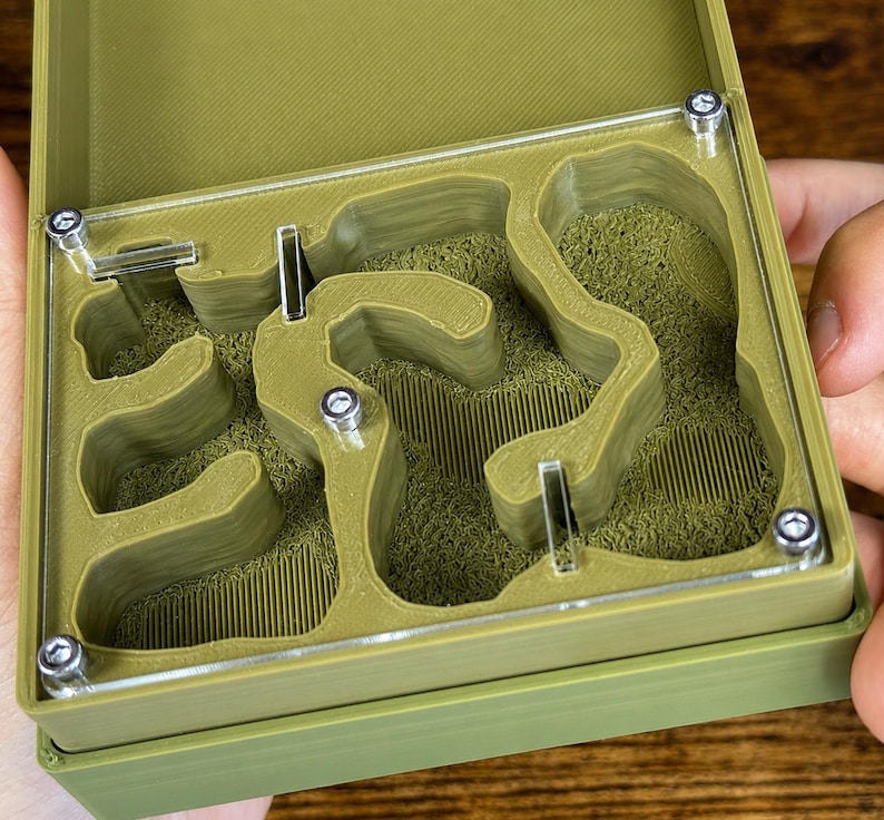 May include: A green ant farm with a clear top, revealing a maze-like interior filled with a textured, green material. The farm is held open, showcasing the design and construction with visible screws.