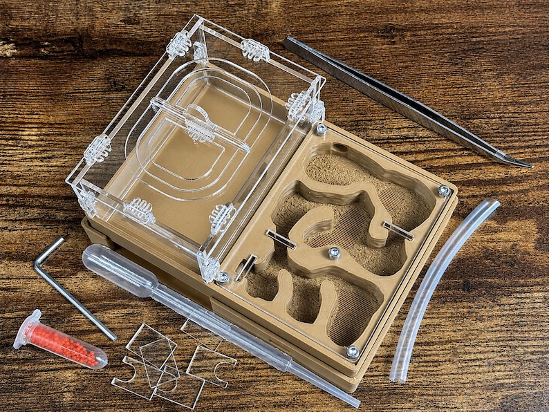 May include: An open ant farm with a clear acrylic top and a tan-colored base, revealing a detailed interior. Accessories include tweezers, a pipette, a small vial with red contents, and clear tubing. The setup is on a wooden surface.