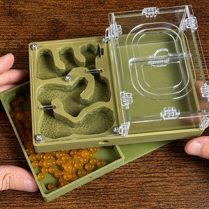 May include: An olive green ant farm with a clear acrylic top. The farm has a detailed interior with tunnels and chambers. A separate tray holds orange, spherical objects. The farm is designed for ant observation and habitat.