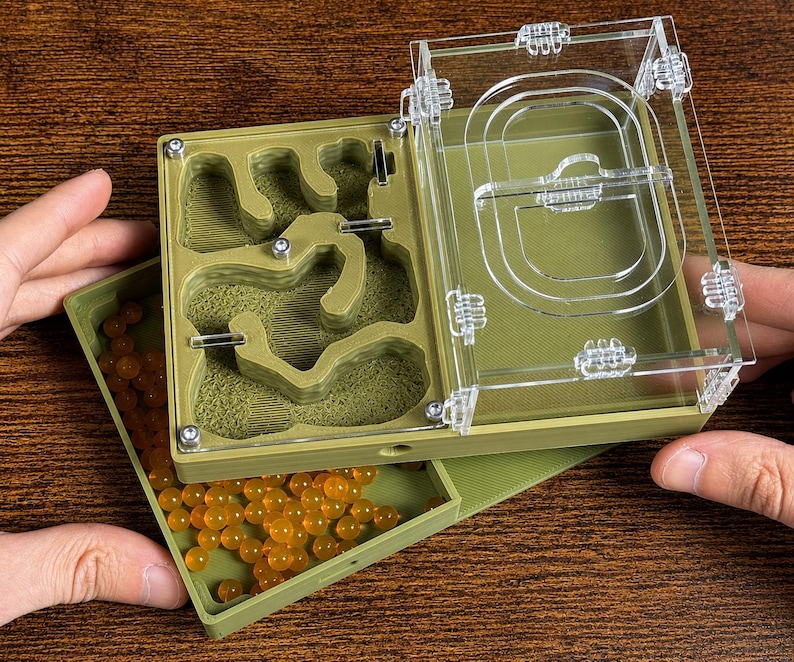 May include: An olive green ant farm with a clear acrylic top. The farm has a detailed interior with tunnels and chambers. A separate tray holds orange, spherical objects. The farm is designed for ant observation and habitat.