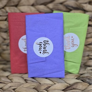 May include: Three wrapped packages in red, purple, and green, each with a white sticker that says "thank you" in a handwritten font. The packages are on a woven wicker surface.