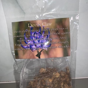 Könnte beinhalten: Ein durchsichtiger Plastikbeutel mit getrockneten braunen Pflanzenteilen und einem Etikett, das eine violette Blume sowie den Text Devils Claw zeigt. Devils claw besitzt magische Schutzeigenschaften gegen Negativität.