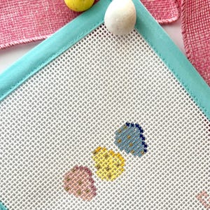 May include: An Easter-themed image featuring a white needlepoint canvas with a turquoise border, adorned with three cross-stitched egg designs in pink, yellow, and blue. Three speckled egg-shaped candies and a pink ribbon complete the composition.