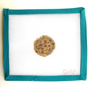 May include: A rectangular white needlepoint canvas featuring a single circular brown and tan cookie design in the center. The canvas edges are bound with a solid teal fabric border, and small pink stitched text is visible in the corner.