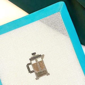 May include: A white needlepoint canvas with a teal border, featuring a cross-stitched image of a French press coffee maker. The canvas has a grid pattern and the initials "G + V" are in the bottom right corner.