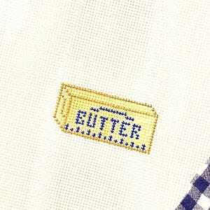 May include: A close-up of a white cross-stitch fabric with a yellow and blue embroidered butter stick design. The word "BUTTER" is stitched in blue. A blue and white checkered pattern is visible in the corner.