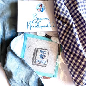 May include: A beginner needlepoint kit featuring a white canvas with a stitched design of a book labeled 901 JOKER, white embroidery floss, a needle, a blue fabric piece, and a blue and white gingham cloth, all packaged with an instructional card.