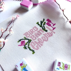 May include: A white fabric square featuring a cross-stitch design with the text "BIRD BATH CHEERS!" in pink, accompanied by green leafy branches and two pink wine glasses. The piece is surrounded by white and pink mahjong tiles and pink ribbon.