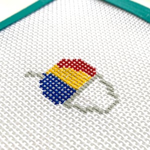 May include: Needlepoint canvas featuring a colorful, cross-stitched design of a beverage with a handle. The drink is composed of red, yellow, and blue sections, with a gray handle. The canvas is stretched on a white background and framed with a teal border.