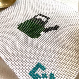 May include: A white needlepoint coaster with a green watering can design and the teal letters "GV". The watering can is detailed with black accents. The coaster is made of a woven material.