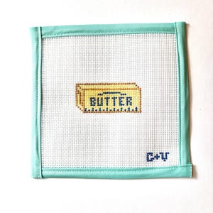 May include: A square, cross-stitched piece featuring a butter graphic. The word "BUTTER" is stitched in dark blue on a yellow rectangle. The piece is framed with a light teal border and has the initials "C+V" in the bottom right corner.