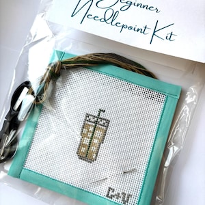 May include: A Beginner Needlepoint Kit in a clear plastic bag. The kit includes a canvas with a stitched iced coffee design, teal fabric border, needles, scissors, and embroidery floss. The text "Beginner Needlepoint Kit" is printed on the top.