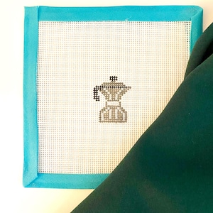 May include: A cross-stitch design featuring a coffee maker, framed with a turquoise fabric border. The coffee maker is in shades of gray and brown, set against a white perforated background. A dark green fabric is draped over the corner.