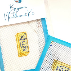 May include: A beginner needlepoint kit featuring a white canvas with a yellow stick of butter design and the word BUTTER stitched in blue. The kit includes a needle and is packaged in a clear plastic sleeve with a header card labeled Beginner Needlepoint Kit.