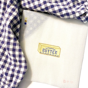 May include: A needlepoint canvas featuring a butter graphic with the word "BUTTER" stitched in blue and gold. The canvas is white and partially covered by a blue and white checkered fabric.