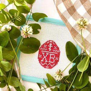 May include: A needlepoint Easter egg design in red and white, framed with a light blue border. The egg is decorated with a cross and the letters XB. The artwork is surrounded by green leaves, white and pink flowers, and a brown and white checkered ribbon.